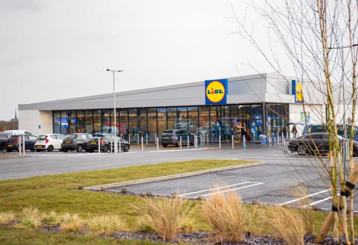 Lidl-Filiale mit Glasfassade, Parkplatz mit mehreren Autos und kahlen Bäumen im Vordergrund