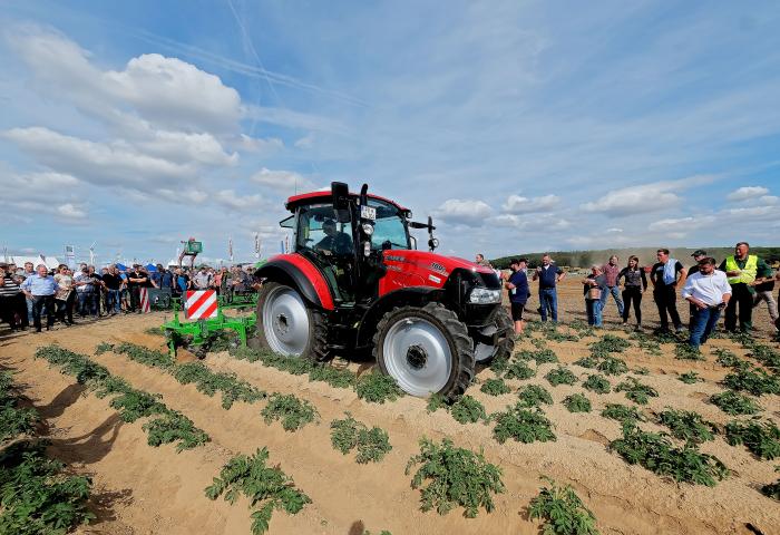 Roter Traktor bei Maschinenvorführung auf Kartoffelfeld, umgeben von Zuschauern unter blauem Himmel