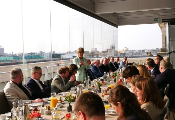 Menschen sitzen an einem langen Tisch auf einer Terrasse mit Blick auf eine Stadt, eine Frau steht und spricht zu der Gruppe während eines Frühstückstreffens.