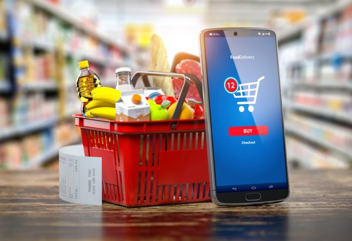 Smartphone mit FoodDelivery-App zeigt Einkaufswagen mit 12 Artikeln und 'Buy'-Button neben rotem Einkaufskorb mit Lebensmitteln und Kassenbon