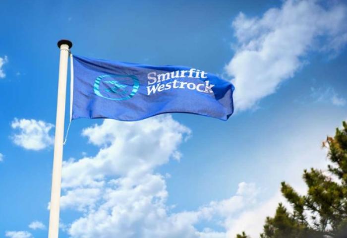 Blaue Flagge mit weißem Schriftzug 'Smurfit Westrock' weht an Fahnenmast vor blauem Himmel mit Wolken