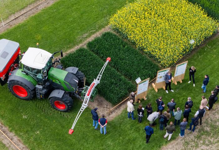 Gruppe von Menschen steht auf einem Feld neben einem grünen Traktor mit Anbaugerät und mehreren Parzellen mit verschiedenen Pflanzen