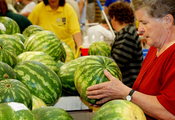 Frau in rotem Oberteil hält eine große Wassermelone auf einem Marktstand mit vielen Wassermelonen