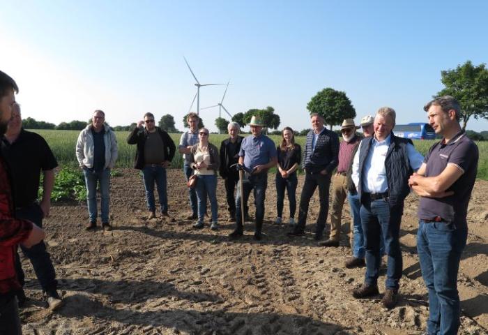 Gruppe von zwölf Personen steht auf einem Acker, im Hintergrund Windräder und Bäume unter blauem Himmel