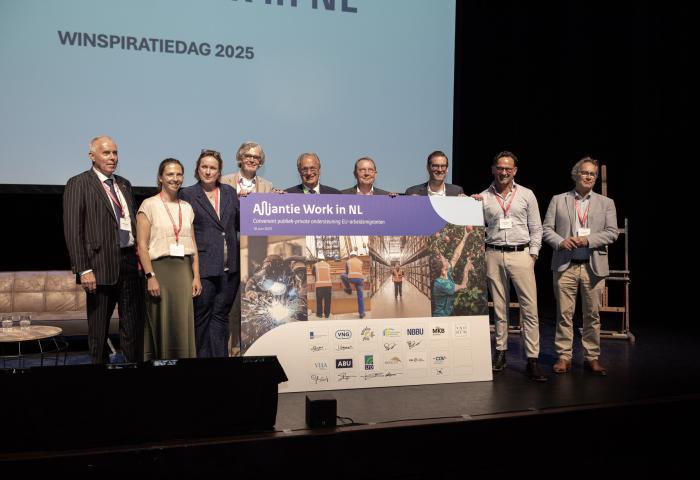 Gruppe von neun Personen auf Bühne halten großes Plakat mit dem Text 'Alliantie Work in NL' und Logos verschiedener Organisationen