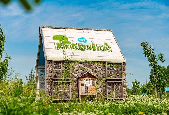 Kleines Haus mit Holzstämmen und Insektenhotels an der Fassade, Schriftzug 'voedselbos' auf dem Dach, umgeben von grüner Wiese und Bäumen