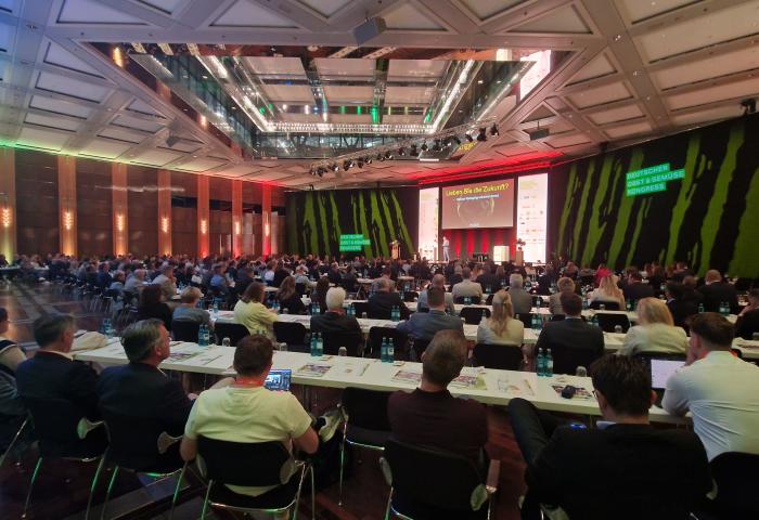 Großer Konferenzsaal mit vielen sitzenden Teilnehmern, Bühne mit Präsentationsbildschirm, grün beleuchtete Wände mit dem Logo "Deutscher Obst und Gemüse Kongress".