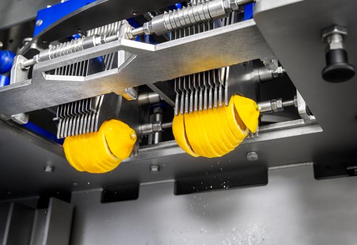 Maschine schneidet zwei Zitronen in dünne Scheiben, die an Metallklingen hängen.