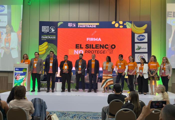 Eine Gruppe von Personen auf einer Bühne, alle tragen orangefarbene T-Shirts mit dem Text 'El Silencio No Protege'. Im Hintergrund sind Fairtrade-Logos und Bananenmuster sichtbar.