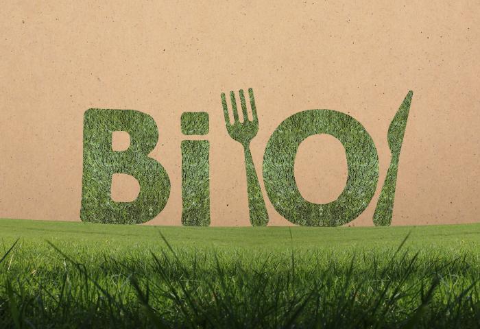 Der Schriftzug 'Bio' ist aus Gras geformt, daneben sind eine Gabel und ein Messer aus Gras dargestellt. Im Vordergrund ist grünes Gras sichtbar.