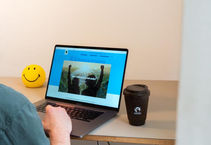 Eine Person sitzt an einem Laptop. Auf dem Bildschirm ist die Fairtrade Kampagnen Website zu sehen Neben dem Laptop steht ein Kaffeebecher mit Fairtrade-Logo und ein gelber Smiley