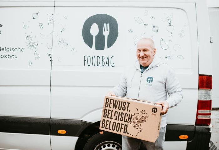 Eine Person hält eine große, braune Box mit dem Text 'BEWUST. BELGISCH. BELOOFD.' vor einem weißen Lieferwagen mit einem grafischen Design von Lebensmitteln und dem Logo 'FOODBAG'.