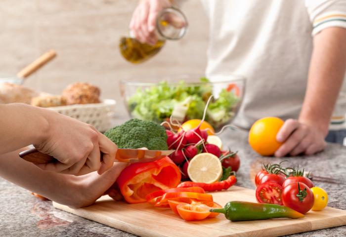 Eine Hand schneidet eine rote Paprika auf einem Holzschneidebrett. Im Hintergrund wird ein Salat in einer Schüssel zubereitet. Weitere Gemüse wie Brokkoli, Tomaten, eine Limette und eine Orange sind sichtbar.