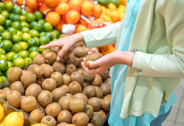 Eine Person hält einen Kiwi in der Hand und steht vor einem Verkaufsstand mit Kiwis, Orangen und Limetten. Die Kiwis sind braun und rundlich.