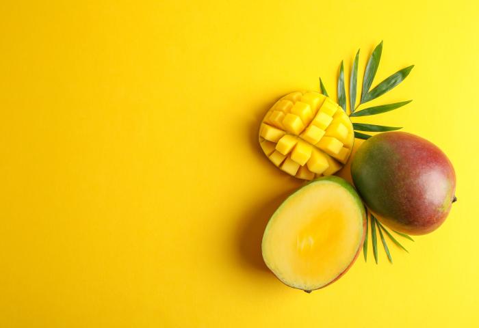 Eine halbierte Mango mit gelbem Fruchtfleisch und eine ganze Mango liegen auf einem gelben Hintergrund, umgeben von grünen Blättern.