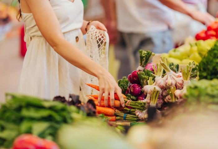 Eine Person in einem weißen Kleid greift nach Karotten auf einem Marktstand, umgeben von frischem Gemüse wie Knoblauch und Blattgemüse.