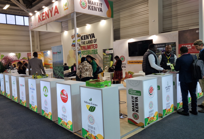 Messestand mit dem Titel 'MAKE IT KENYA'. Tische mit Logos von verschiedenen Unternehmen. Menschen interagieren am Stand.