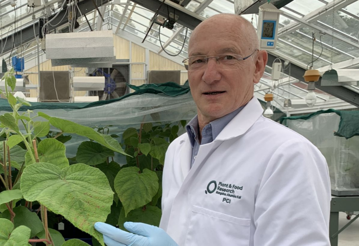 Dr. Joel Vanneste steht in einem Gewächshaus, trägt Arbeitskleidung. Im Hintergrund sind Pflanzen und Laborausrüstung sichtbar.