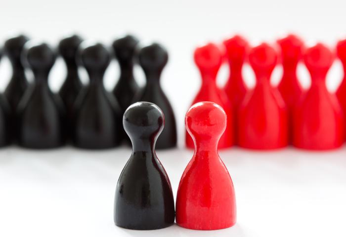 Zwei Spielfiguren, eine schwarz und eine rot, stehen eng beieinander im Vordergrund. Im Hintergrund sind mehrere schwarze und rote Spielfiguren in Gruppen angeordnet.