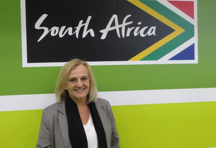 Retha Louw steht vor einer Wand mit dem Schriftzug 'South Africa' und dem südafrikanischen Flaggenmuster in den Farben Grün, Gelb, Blau und Rot.