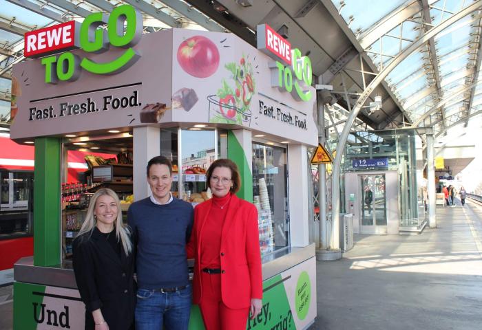 Helen Schnürle, Michael Mayer-Sonnenburg und Jenny Ramme stehen vor einem REWE To Go Kiosk mit Obst- und Lebensmittelangeboten. Der Kiosk ist farbenfroh gestaltet und befindet sich in einem Bahnhof.