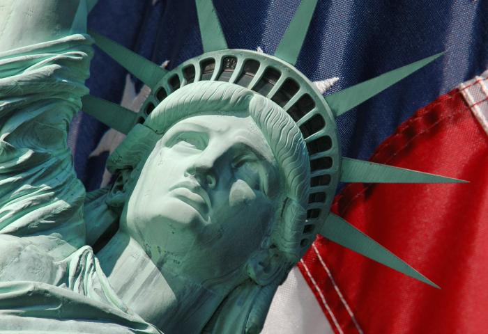 Die Freiheitsstatue mit grünem Kupferfinish, Blick nach oben, vor einem Hintergrund der amerikanischen Flagge mit roten, weißen und blauen Streifen und weißen Sternen.
