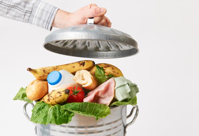 Eine Hand hebt den Deckel eines Metallmüllbehälters, der mit Lebensmitteln gefüllt ist, darunter Bananen, ein Brot, Milch, eine Tomate, Schinken und Salat.