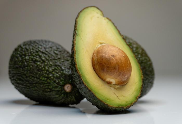 Eine aufgeschnittene Avocado mit einem großen, braunen Kern in der Mitte. Im Hintergrund sind zwei ganze Avocados sichtbar.