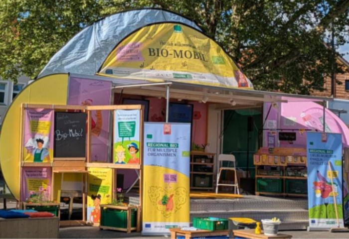 in mobiler Informationsstand mit bunten Wänden in Gelb und Pink, ausgestattet mit Plakaten und einem kleinen Tisch. Auf einer Tafel steht 'Bio-Mobil!'.