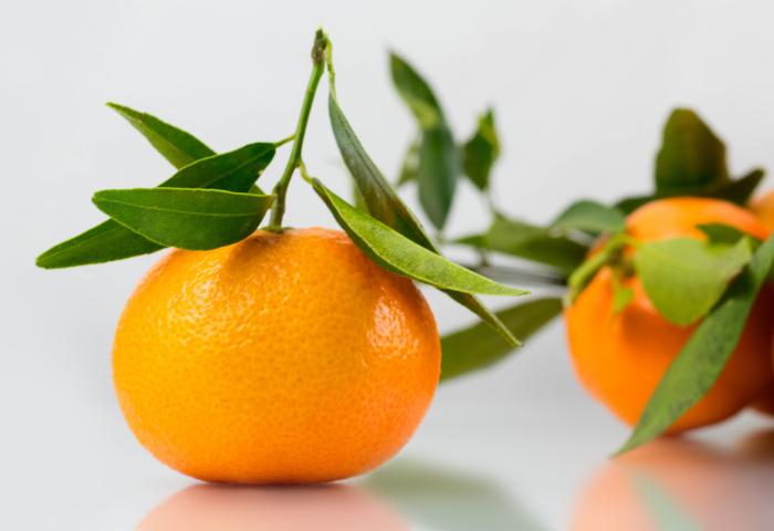 Eine Clementine mit grünem Blatt am Stiel, daneben unscharf weitere Clementinen mit Blättern auf hellem Untergrund