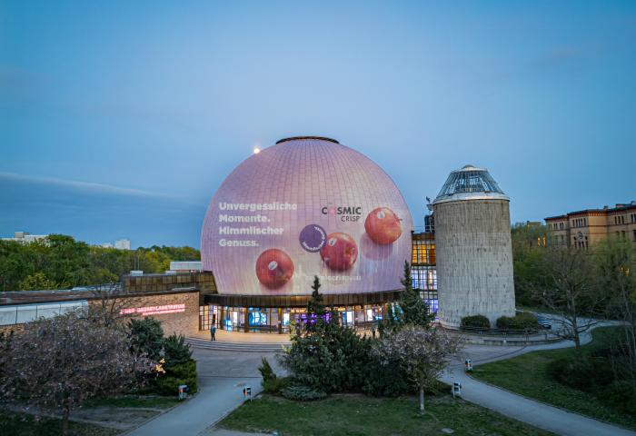 Das Berliner Planetarium mit einer großen Projektion von Cosmic Crisp® Äpfeln auf der Kuppel. Text: 'Unvergessliche Momente. Himmlischer Genuss.'