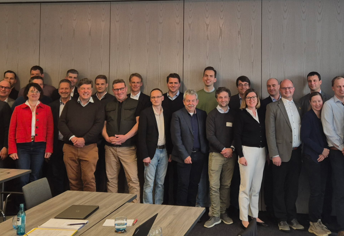 Gruppenfoto von vielen Personen in einem Konferenzraum. Tische mit Unterlagen und Wasserflasche im Vordergrund.