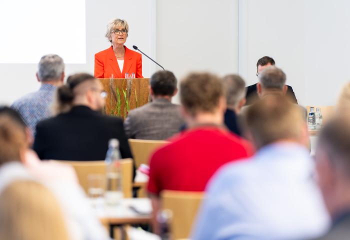 Eva Kähler-Theuerkauf spricht an einem Rednerpult vor einem Publikum. Das Publikum sitzt in Reihen an langen Tischen, auf einem ist auch eine Wasserflasche zu erkennen.