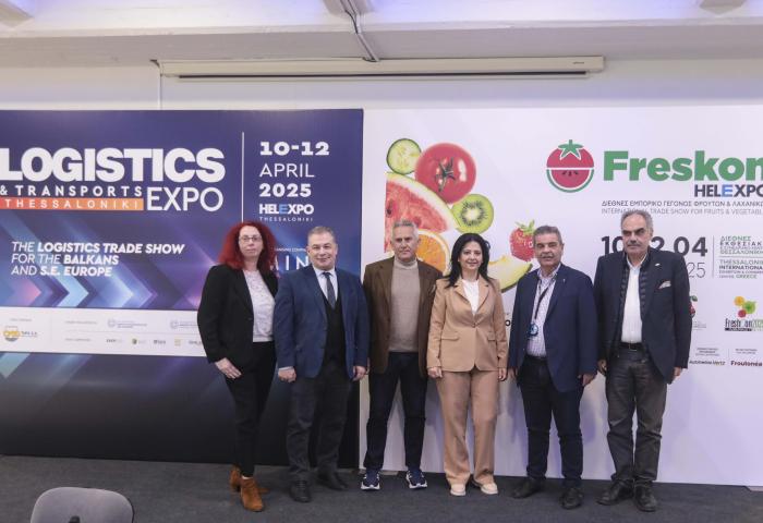 Sechs Personen stehen in einer Reihe vor einer Wand mit zwei Plakaten. Links ein Plakat mit dem Text 'LOGISTICS & TRANSPORTS EXPO'. Rechts ein Plakat mit dem Logo 'Freskon' und dem Datum '10-12 APRIL 2025', sowie Bildern von Obst.