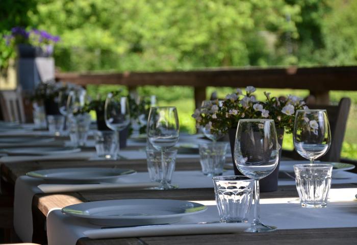Langer Holztisch im Freien mit weißem Tischtuch, gedeckt mit weißen Tellern, Weingläsern, Wassergläsern und Blumentöpfen mit weißen und lila Blumen.