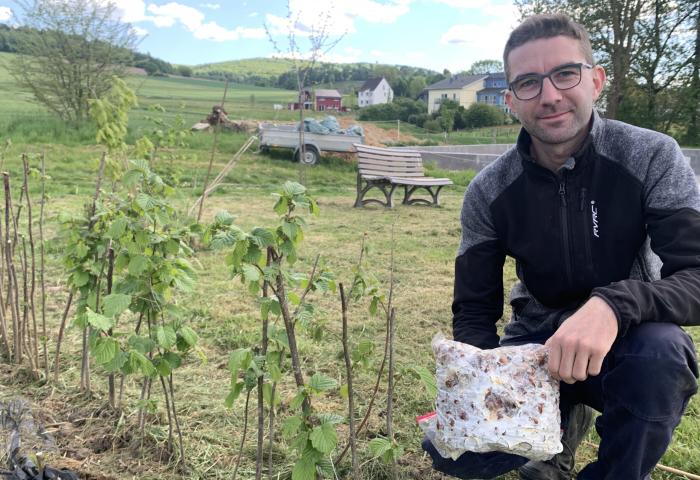 Hendrik Hoffmann kniet im Freien und hält ein Paket mit Pflanzenmaterial in der Hand. Im Hintergrund sind Sträucher und ein Wohnhaus sichtbar.