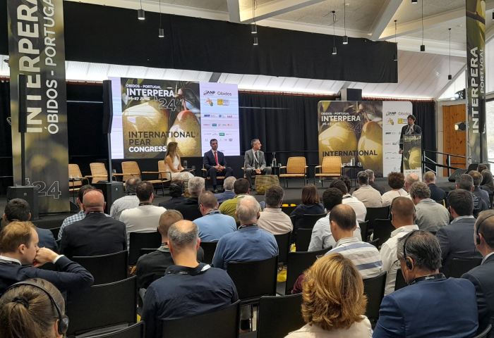 Blick auf eine Bühne mit mehreren Personen im Publikum. Auf der Bühne sitzen drei Personen und eine weitere steht rechts am Rand und spricht am Mikrofon. Im Hintergrund ein Banner mit 'INTERPERA 24 INTERNATIONAL PEAR CONGRESS'.