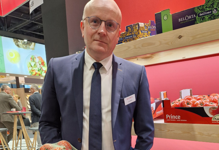 Jo Lambrecht hält ein Netz mit Tomaten, steht vor einem Regal mit weiteren Tomatenkartons bei einem Messestand.