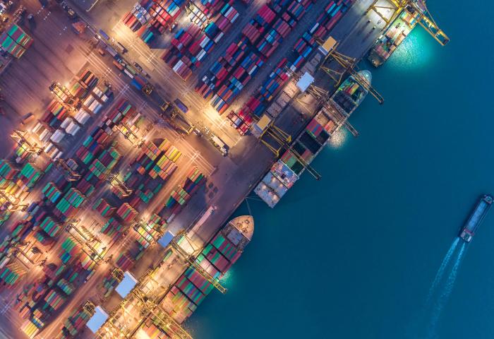 Luftaufnahme eines beleuchteten Containerhafens mit zahlreichen bunten Containern und einem Schiff, das eine Wasserstraße entlangfährt.