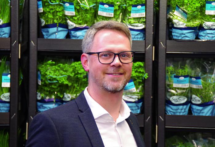 Nils Doerwald steht vor einem Regal mit frischen Kräutern in Töpfen, die in blauen Verpackungen von 'Sangrar' präsentiert werden.