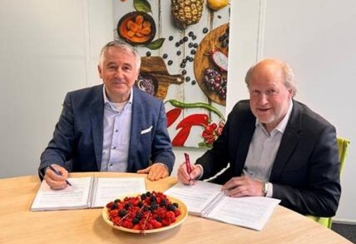 Richard Schouten und René van der Burg sitzen an einem Tisch und unterschreiben Dokumente. Vor ihnen steht eine Schüssel mit Beeren.