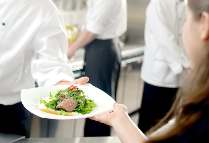 Ein Koch in einer weißen Kochjacke reicht einen Teller mit Fleisch und Salat an eine Person. Im Hintergrund sind weitere Köche zu sehen.