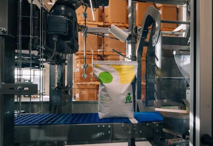 Eine Maschine verpackt einen weißen Sack mit grünem und gelbem Logo von Rijk Zwaan. Im Hintergrund sind braune Kartons sichtbar.