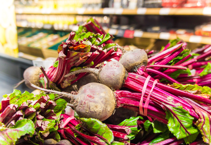 Bund frischer Rote Beete mit grünen Blättern, violetten Stielen und erdigen Knollen, in einem Supermarktregal.