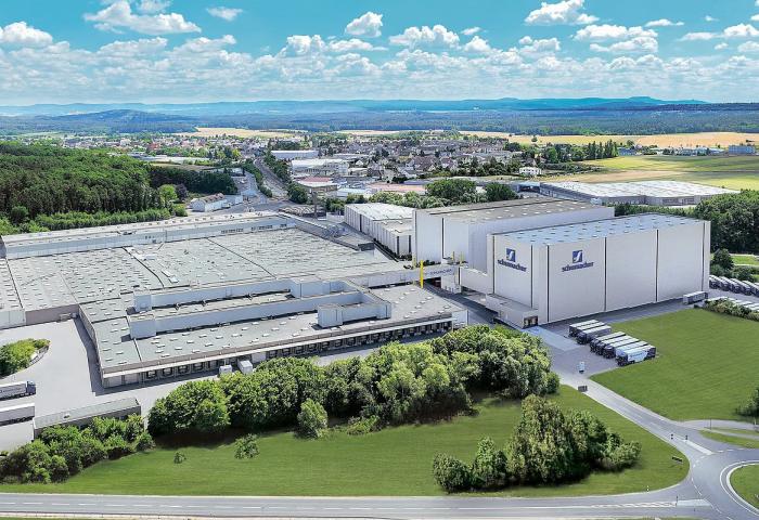 Luftaufnahme der Schumacher Packaging-Werke mit mehreren Dächern, umgeben von Wäldern und Feldern. Im Vordergrund sind Lkw und eine Straße sichtbar.