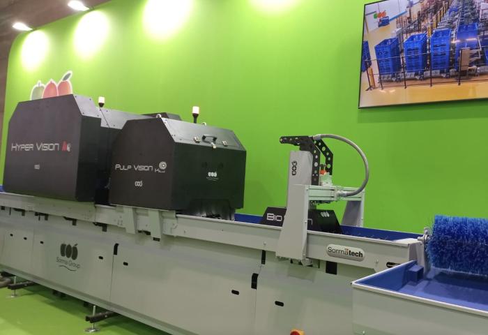 Maschine zur Fruchtsortierung mit zwei großen schwarzen Geräten, beschriftet mit 'HYPER VISION' und 'PULP VISION', auf einem Förderband. Im Hintergrund ein Bildschirm mit blauen Kisten.