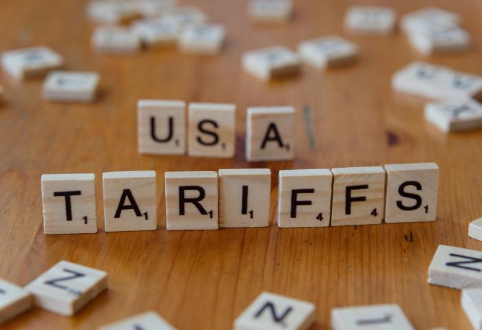 Holzbuchstaben auf einem Tisch bilden das Wort 'TARIFFS' und 'USA'. Die Buchstaben sind in einem Scrabble-ähnlichen Stil angeordnet.