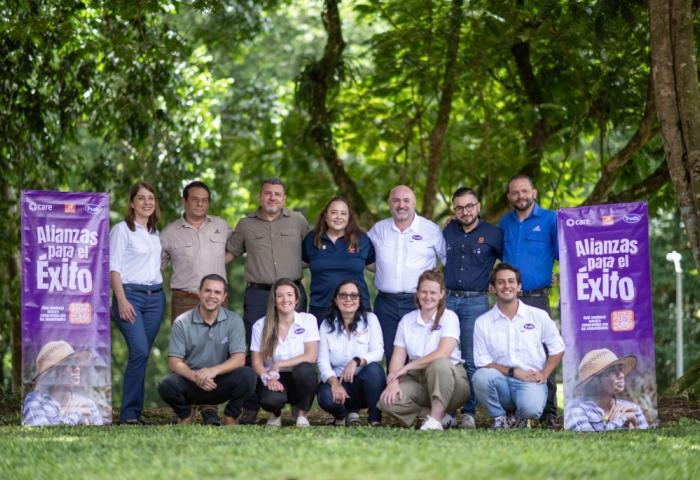 Gruppenfoto von 12 Personen im Freien, umgeben von Bäumen. Im Hintergrund zwei Banner mit dem Text 'Alianzas para el Éxito'.