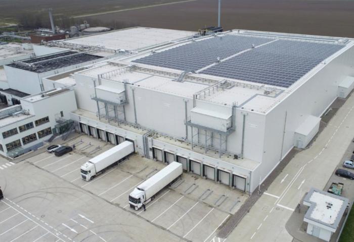 Luftaufnahme eines großen Lagergebäudes mit Solarpanelen auf dem Dach. Vor dem Gebäude stehen mehrere Lkw an den Ladebuchten.