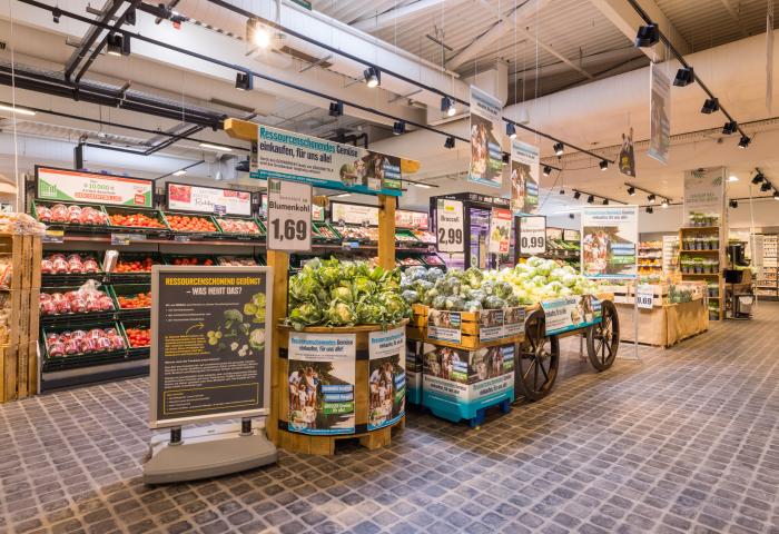 Verkaufsfläche mit Regalen und Wagen voller Blumenkohl, Preisschilder und Informationsplakate in einem Supermarkt.